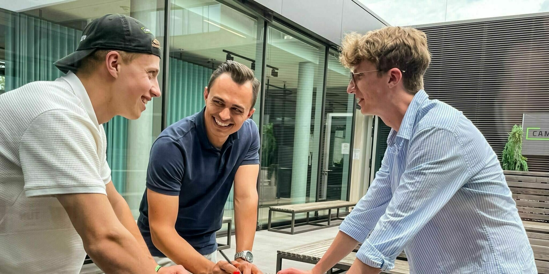 Three men standing around a round table, laughing, looking at each other.